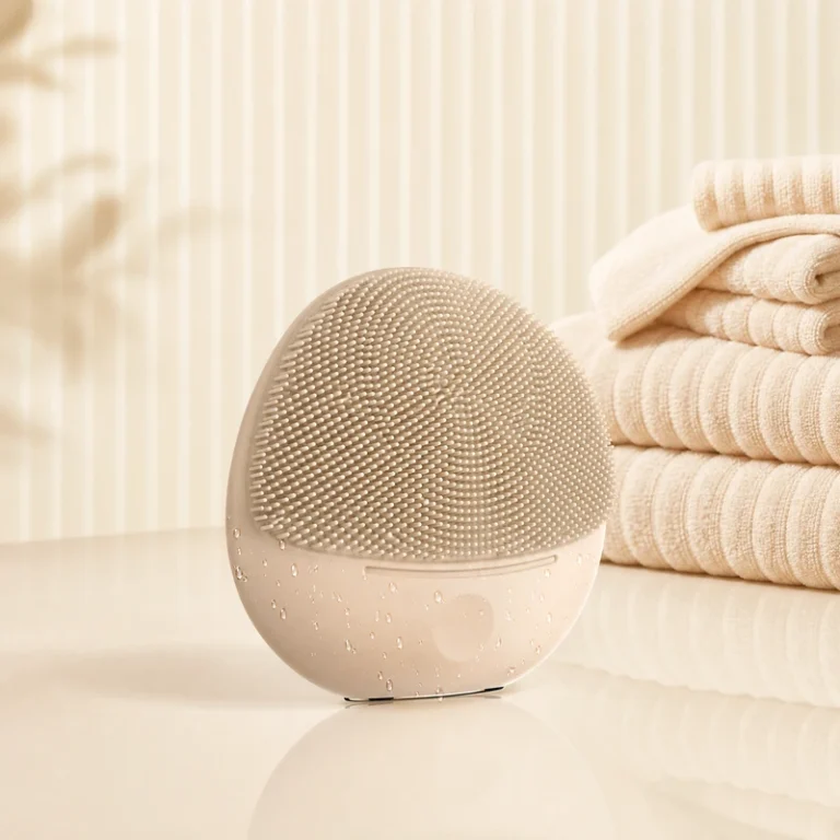 Champagne-gold silicone facial cleansing device with textured brush head and water droplets on a luxury vanity.
